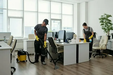 Two professional cleaners from 'Eco Techno Clean' cleaning a bright, modern office space: one vacuuming a chair and the other sanitizing a desk.